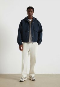Veste légère bleu marine avec capuche, zip avant et grandes poches. Portée sur une chemise crème et assortie avec un pantalon clair et des baskets grises.