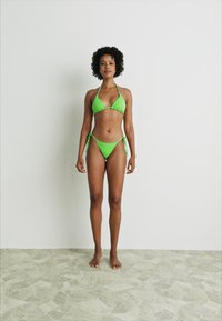 Lys grønn bikini med trekantet topp og lav skjæring, med knyte-detaljer i sidene. Glatt tekstur, minimalistisk design.