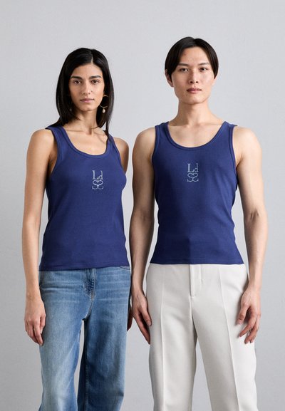 Marineblå tanktopper med ribbet tekstur som har et sentralt logo. En modell har på seg lyseblå jeans, mens den andre har på seg hvite skræddersydde bukser.