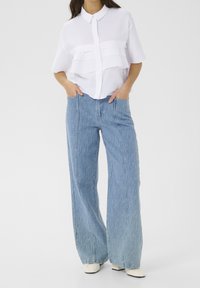 Kvindelig iført løs, lys blå jeans og en textureret hvid kortærmet bluse, stående med hænderne i lommerne på en ensfarvet baggrund.