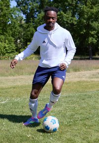 Atleta con sudadera blanca y pantalones cortos azul marino, regateando un balón de fútbol azul y blanco sobre césped, con zapatos de fútbol de colores rosa y verde brillante.