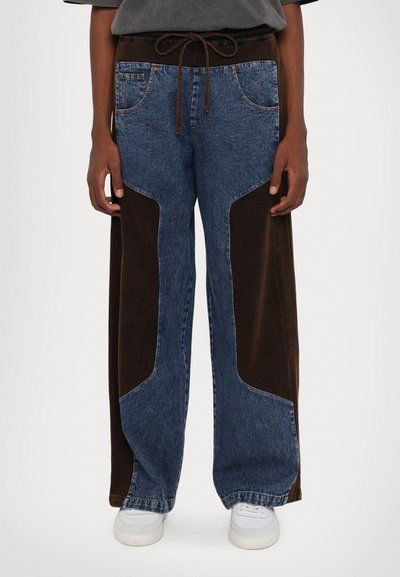Des pantalons larges en denim et en velours côtelé avec des panneaux latéraux marron foncé, une taille avec cordon de serrage marron et des poches avant, complétés par des baskets blanches.