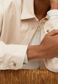 Osoba w beżowej kurtce na białym swetrze z ribbingiem, opierająca ramię na plecionym koszu.