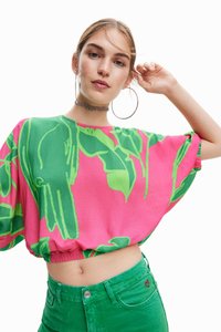 Lys pink cropped top med grønne bladmønstre, løs pasform, rund halsudskæring og elastisk kant. Model iført grønne højtaljede shorts.