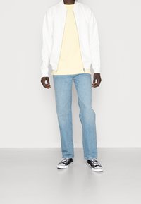 Vit bomberjacka över en ljusgul t-shirt, tillsammans med blekta blå jeans och svart-vita sneakers. Rent utförande och avslappnad passform.