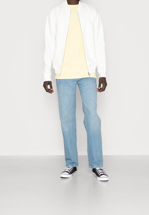 Chaqueta bomber blanca sobre una camiseta ligera amarilla, combinada con jeans desgastados de color azul y zapatillas en blanco y negro. Líneas limpias y ajuste relajado.