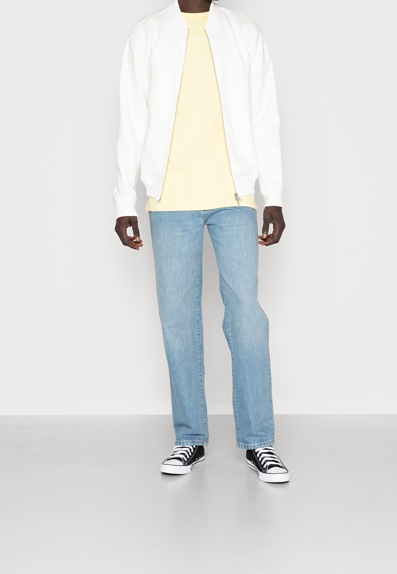 Vit bomberjacka över en ljusgul t-shirt, tillsammans med blekta blå jeans och svart-vita sneakers. Rent utförande och avslappnad passform.