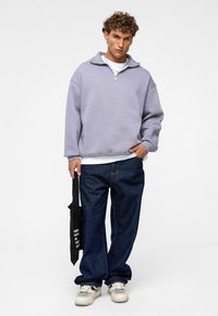 Lavendel rits-sweater, ontspannen pasvorm, met verlaagde schouders. Gecombineerd met donkere denimjeans en sneakers met witte zolen. Inclusief zwarte schoudertas.