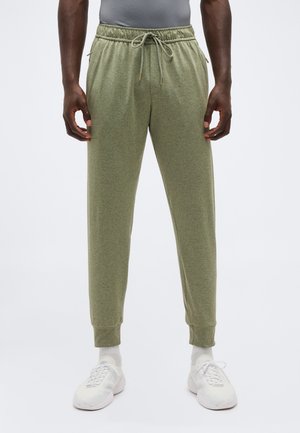 Muž nosiaci olivovo zelené teplákové nohavice s elastickým pásom a šnúrkou, biele ponožky a biele tenisky na jednoduchom pozadí.