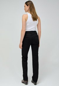 Jeans neri, aderenti con vita alta e gambe svasate. Le tasche posteriori presentano cuciture sottili. Il modello indossa un top bianco e scarpe marroni.