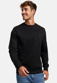 Felpa nera in misto cotone, con scollo rotondo, maniche lunghe e un piccolo logo sul petto; abbinata a jeans blu.