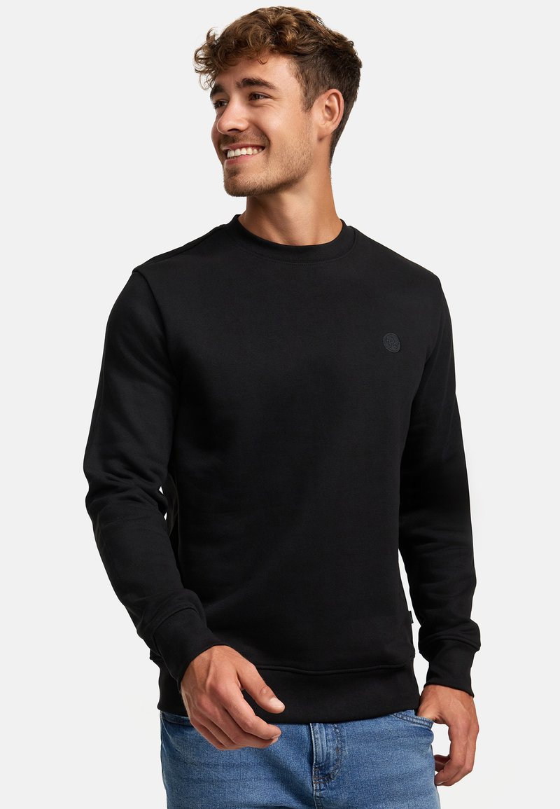Felpa nera in misto cotone, con scollo rotondo, maniche lunghe e un piccolo logo sul petto; abbinata a jeans blu.