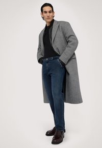 Šedý vlněný kabát s přizpůsobeným, oversized designem, černý rolák, tmavě modré džíny a tmavě hnědé mokasíny, vystavené na neutrálním pozadí.