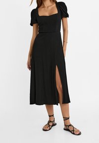 Robe midi noire avec un décolleté carré, manches bouffantes, tissu texturé et fente latérale. Assortie à des sandales noires à brides.