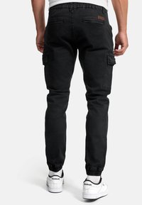 Bărbat purtând pantaloni cargo negri cu manșete elastice și adidași albi Reebok, stând cu spatele la fundal alb.