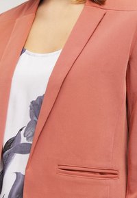 Marynarka w odcieniu różowego bladoróżowego, o gładkiej fakturze, dopasowanym kroju i pojedynczej kieszeni na piersi; noszona na bluzce w kwiatowy wzór.