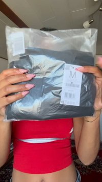 Schwarzes Kleidungsstück in transparenter Plastikverpackung mit sichtbarem Größenetikett (M) und Barcode. Die Verpackung hat eine glatte Oberfläche.