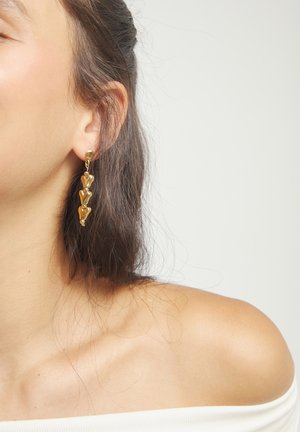 Primer plano de la oreja izquierda de una mujer que lleva pendientes colgantes de oro en forma de corazones apilados, con el hombro descubierto y el cabello oscuro recogido hacia atrás.