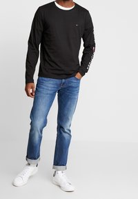 Schwarzes Langarm-T-Shirt mit weißem Kragen und Markenlogo auf der linken Brust und dem Ärmel, kombiniert mit blauen Jeans und weißen Sneakers.
