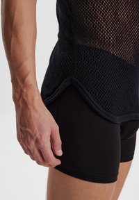 Sort mesh tank top med et struktureret design, parret med sorte tætsiddende shorts. Synlig hånd hviler langs shorts' kanten.