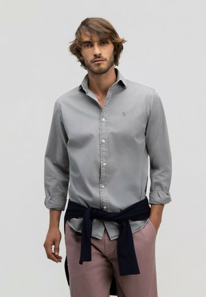 Hombre con cabello de longitud media que lleva una camisa abotonada gris claro, pantalones rosas y un suéter oscuro atado alrededor de su cintura.