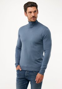 Přiléhavý svetřík s rolákem v jemně modré barvě, vyrobený z hladkého pleteného materiálu. Má dlouhé rukávy a decentní branding na hrudi. Kombinován s tmavými džíny.
