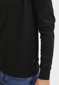 Maglione nero lavorato a maglia con maniche lunghe, polsini e orlo a costine. Presenta un piccolo etichetta sul lato della cucitura. Il materiale sembra morbido e liscio.