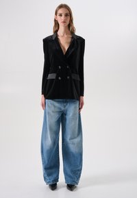 Zwart fluwelen double-breasted blazer met satijnen accenten en lange mouwen, gecombineerd met losse, lichtblauwe spijkerbroeken en zwarte puntige hakken.