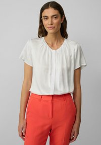 Weiße kurzärmlige Bluse mit rundem Ausschnitt und gefaltetem Vorderteil, kombiniert mit korallenfarbenen, figurbetonten Hosen. Glattes, leichtes Material.