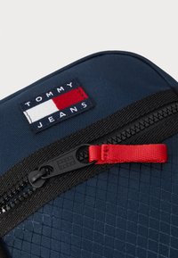 Pochette di colore blu navy con texture, dotata di una zip rossa e accessori neri. Presenta un'etichetta ricamata rettangolare con il logo "TOMMY JEANS."