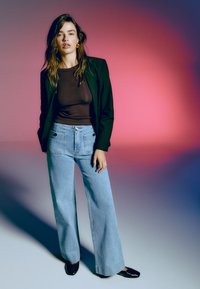 Žena s dlhými vlasmi má na sebe čierny blazer, hnedý priliehavý top, svetlomodré široké nohavice a čierne topánky pred pozadím v odtieňoch od ružovej po fialovú.