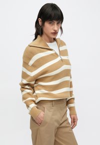Prugasti pulover u smeđoj i bijeloj boji, s ovratnikom na patentni zatvarač i rebrastom teksturom. Uparen s bež hlačama.