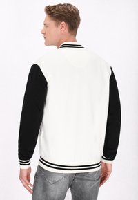 Jachetă varsity albă și neagră cu manșete și tiv în dungi ribbed, având mâneci negre și o textură netedă.