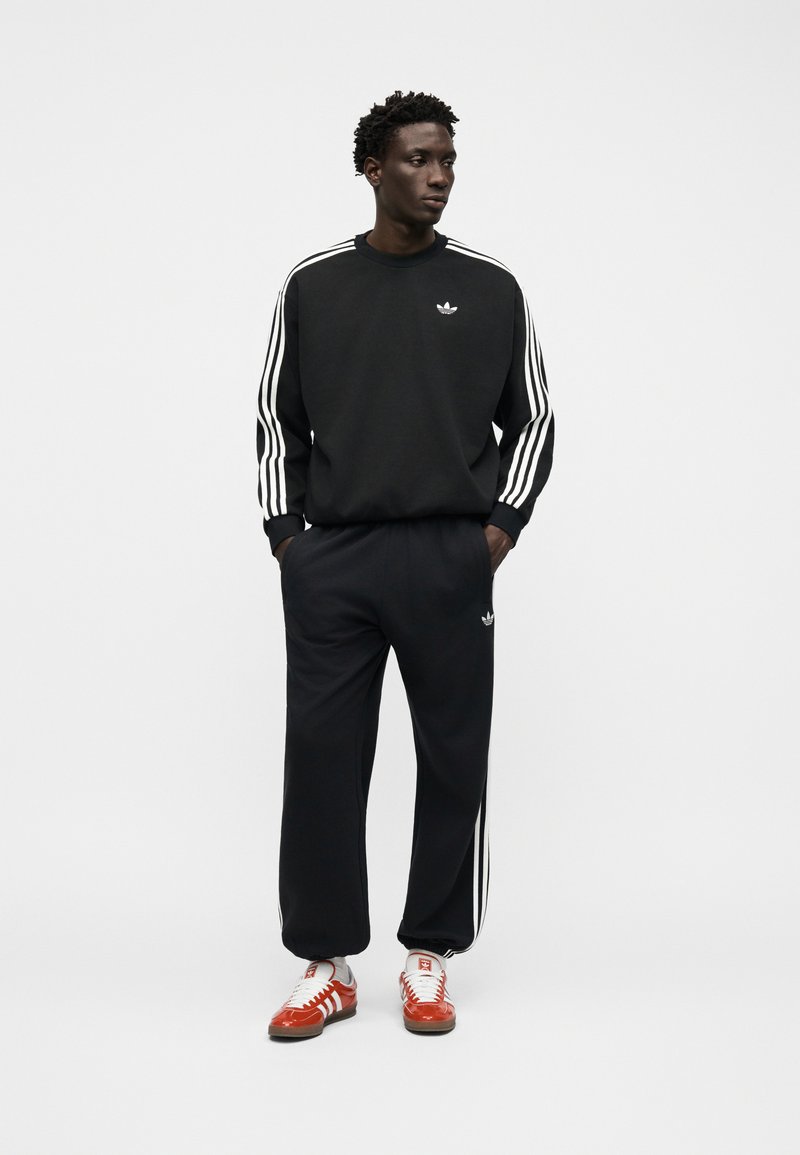 Man wearing black Adidas tracksuit with white stripes and logos, standing with hands in pockets, looking to the side, on plain white background.