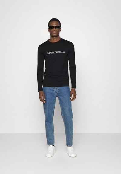Juodas ilgos rankovės marškinėlis su "EMPORIO ARMANI" logotipu, derinamas su šviesiai mėlynomis džinsinėmis kelnėmis ir baltais sportiniais bateliais; modelis nešioja tamsius akinius nuo saulės.