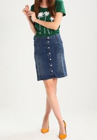 Denimhame, jossa on etuknappi, kaksi sivutaskua ja kynsilinja; yhdistetty vihreän graafisen t-paidan ja keltaisten korkokenkien kanssa.