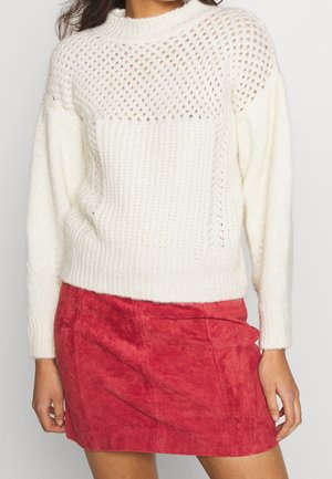Femme portant un pull en maille blanc avec des détails en ajouré, associé à une mini-jupe en suède rouge, debout devant un fond uni.