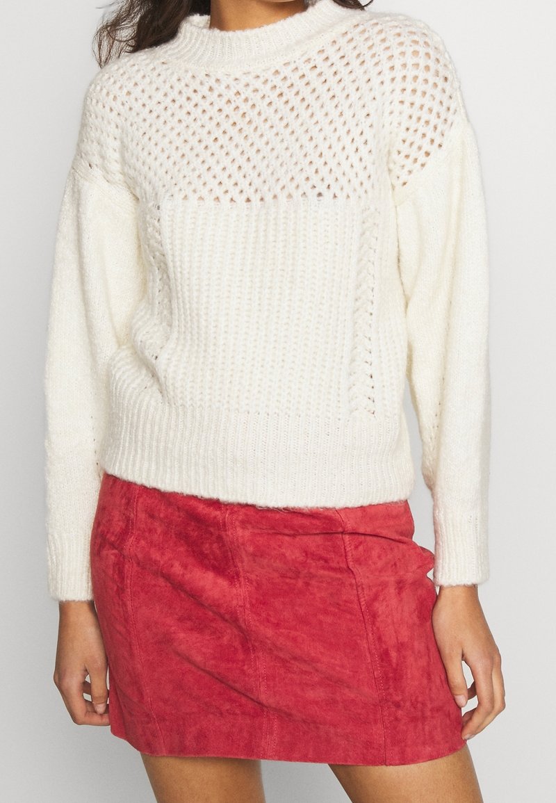 Femme portant un pull en maille blanc avec des détails en ajouré, associé à une mini-jupe en suède rouge, debout devant un fond uni.