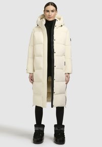 Puffy, knee-length coat in off-white, featuring a hood, snap buttons, and side pockets. Paired with black, insulated boots.