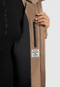 Beige yderjakke med en glat tekstur, der har en sort inderside og en lynlåslomme. Etiketten indeni læser "Et varmt kram lavet af stof."