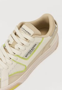 Beige Sneaker mit grünen Akzenten, Lochmuster und weißen Schnürsenkeln. Verfügt über einen gepolsterten Schaft und eine strukturierte Sohle. Logo auf der Zunge angezeigt.