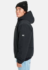 Jeune homme portant une veste noire à capuche, un bonnet noir et un jean bleu clair, se tenant de profil sur fond blanc.