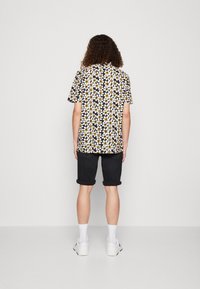 Camicia a maniche corte con sfondo nero e motivi floreali bianchi e gialli, abbinata a pantaloni corti scuri arrotolati e sneakers bianche.