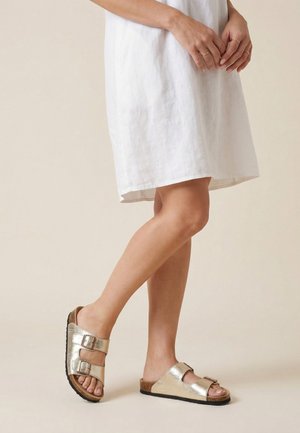 Kvinde iført hvid kjole og metallisk gyldne sandaler med dobbelt spænde stående med det ene ben let bøjet på beige baggrund.