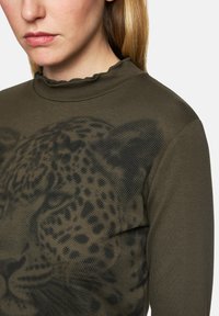 Olive green long-sleeve top with a high neck and ruffled trim, featuring a large black leopard print design across the front.