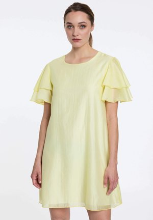 Vrouw met lichtbruin haar in een staart, gekleed in een lichtgele korte mouw jurk met gelaagde zwierige mouwen, tegen een witte achtergrond.