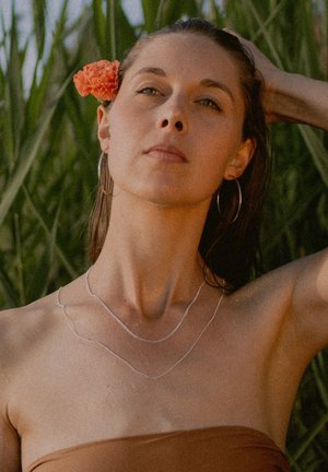 Silberne doppelte Halskette, die auf einer nackten Schulter liegt; der Hintergrund zeigt grünes Laub, mit einer korallenfarbenen Blume im Haar.