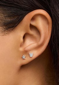Pendientes de plata en forma de estrella y corona, con un acabado suave y brillante. Llevados en el lóbulo de una oreja con cabello oscuro parcialmente visible.