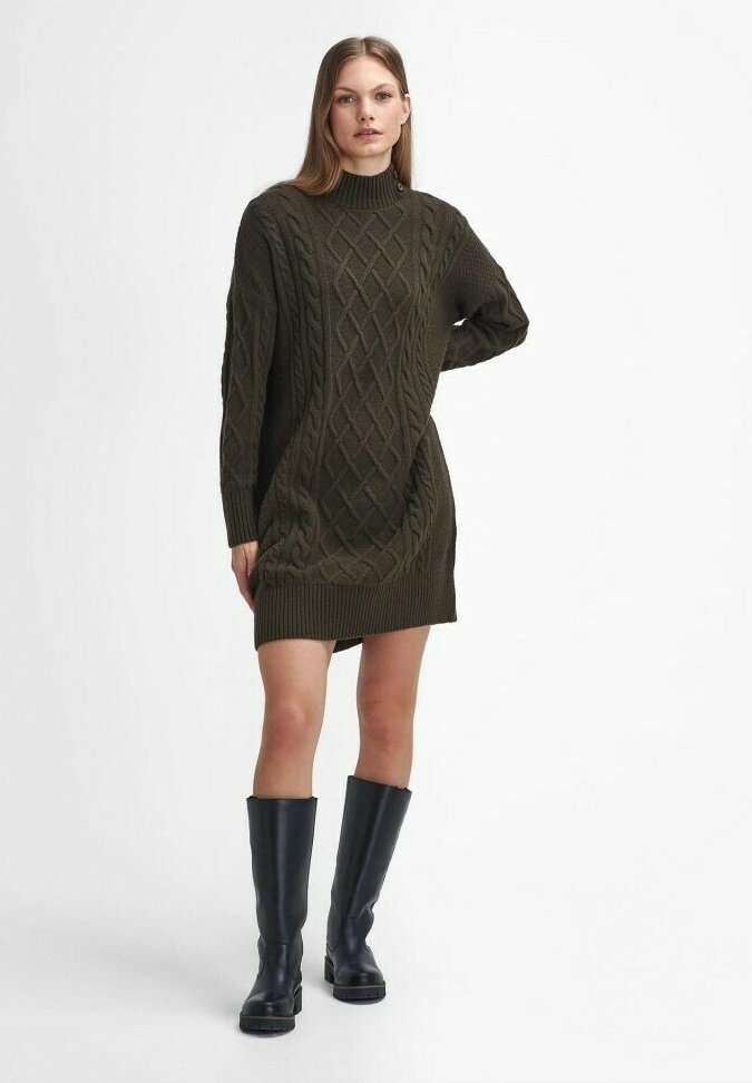 Nainen, jolla on päällään tummanvihreä palmikkoneulemekko ja mustat polvimittaiset saappaat, seisoo käsi lanteellaan valkoista taustaa vasten.