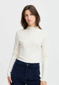 Top de manga larga de canalé color crema con un cuello alto y rizado, y una silueta ajustada, combinado con pantalones de pana azul marino que tienen bolsillos traseros.